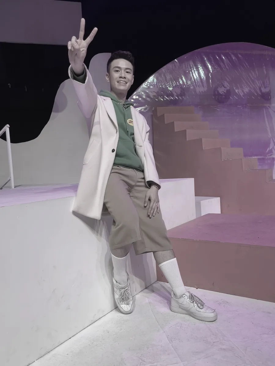 舞台布景前，穿浅色大衣比出手势